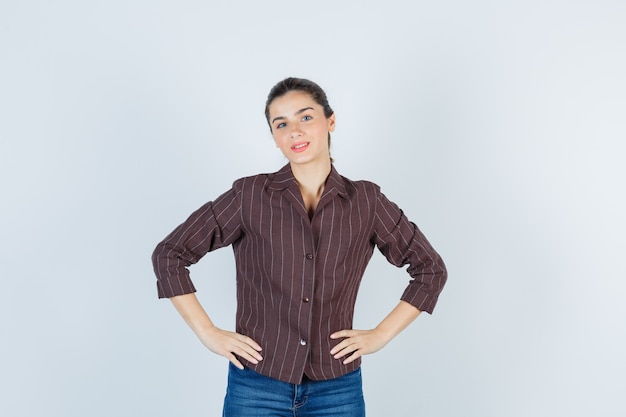 mujer manteniendo las manos en la cintura en camisa, jeans y mirando confiada, vista frontal.