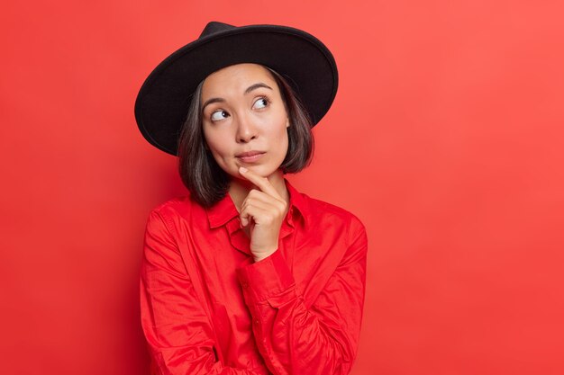 La mujer mantiene la barbilla con la mano en pensamientos profundos, considera que algo viste una camiseta de moda hatt negra posa en rojo