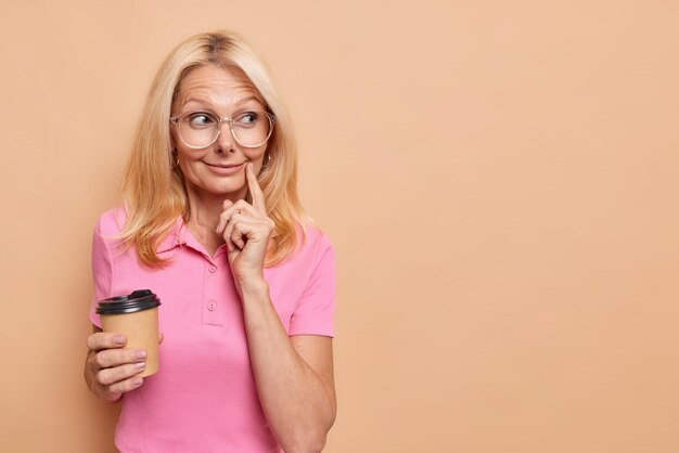 La mujer mantiene el dedo índice cerca de la esquina de los labios mira hacia otro lado con una sonrisa suave disfruta de un café aromático viste una camiseta rosa informal aislada en beige