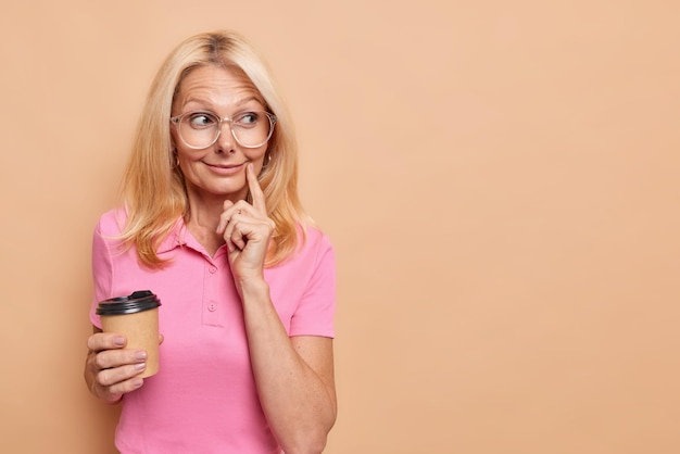 La mujer mantiene el dedo índice cerca de la esquina de los labios mira hacia otro lado con una sonrisa suave disfruta de un café aromático viste una camiseta rosa informal aislada en beige