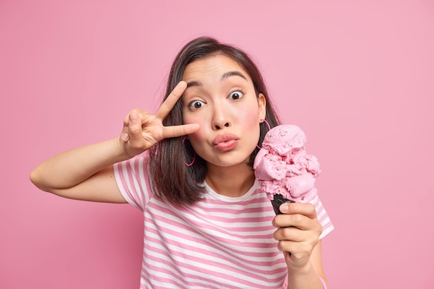 La mujer mantiene los labios doblados hace un gesto de paz sobre el ojo vestido con una camiseta a rayas sostiene un delicioso helado para los golosos