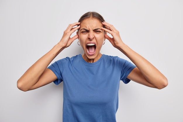 la mujer mantiene las manos en la cabeza grita enojada mantiene la boca bien abierta pierde el control tiene un colapso mental grita furioso viste una camiseta azul sobre blanco alivia del estrés