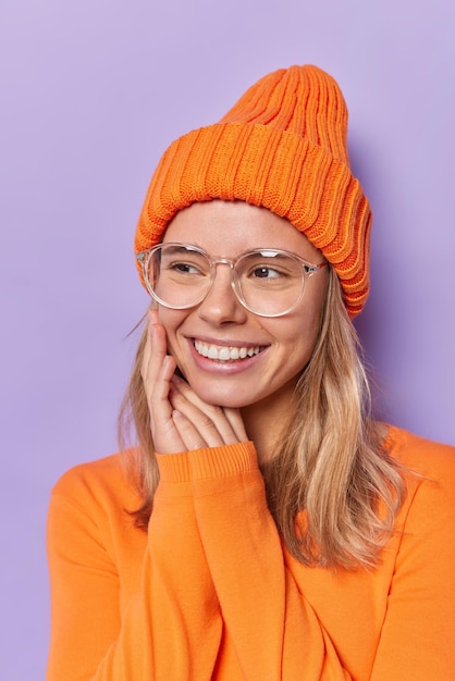 la mujer mantiene las manos cerca de la cara sonríe suavemente con los dientes mira hacia otro lado viste un jersey de manga larga naranja y un sombrero aislado en morado tiene pensamientos agradables. Emociones felices