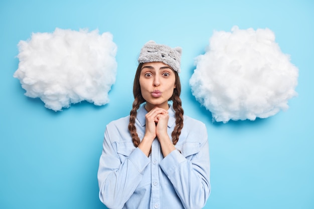 La mujer mantiene las manos debajo de la barbilla, mantiene los labios doblados, lleva los ojos vendados y la camisa informal se despierta después de un sueño saludable aislado en azul