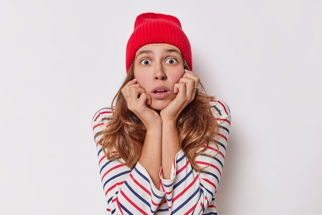 La mujer mantiene las manos debajo de la barbilla tiene una expresión asustada conmocionada escucha algo con aguantar la respiración usa un sombrero rojo y un jersey a rayas aislado en blanco oye noticias asombradas