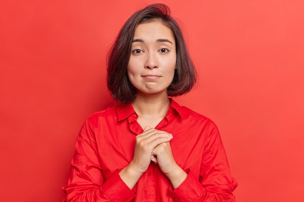 La mujer mantiene las manos juntas en gesto de oración tiene expresión de lástima pide disculpa se ve con gran esperanza en la cámara usa poses de camisa en rojo brillante