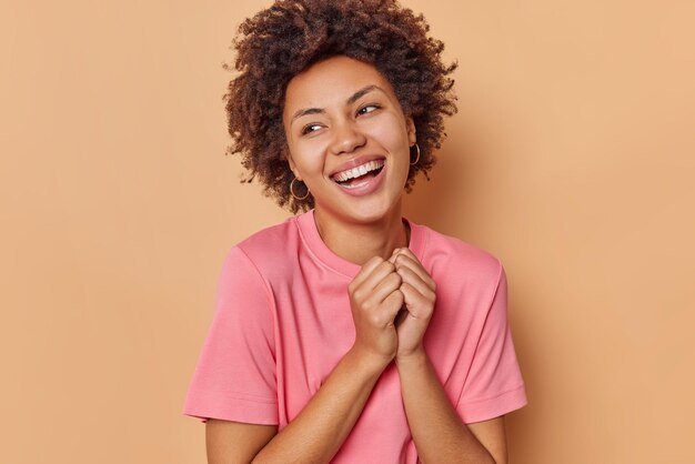 la mujer mantiene las manos juntas sonríe ampliamente y mira hacia otro lado expresa emociones positivas viste una camiseta casual rosa aislada en beige admira algo agradable.