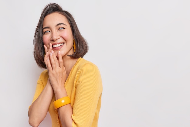 La mujer mantiene las manos juntas sonríe con los dientes concentrados con expresión de alegría se siente muy contenta de usar un jersey amarillo y poses de brazalete en un espacio en blanco blanco