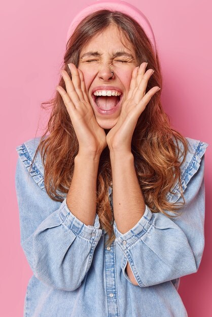 mujer mantiene las manos en las mejillas exclama en voz alta de felicidad mantiene los ojos cerrados viste camisa de mezclilla y diadema posa contra rosa expresa emoción