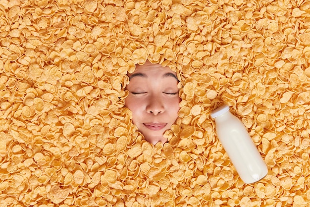 Foto gratuita la mujer mantiene los ojos cerrados imagina cómo toma un delicioso desayuno enterrado en copos de maíz muestra solo rostro va a comer cereales con leche prefiere productos saludables. concepto de nutrición