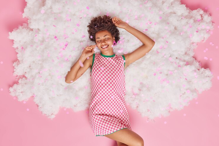 Foto gratuito la mujer mantiene los ojos cerrados por el placer viste un vestido disfruta de la libertad y la relajación posa en una nube blanca con confeti volador alrededor de rosa