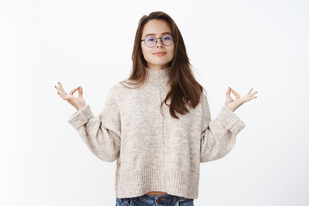 La mujer mantiene la paciencia, está tranquila y en paz mientras posa con suéter y gafas mirando al frente, sonriendo de pie en posición de loto con gesto de mudra, meditando o haciendo yoga para relajar la mente.