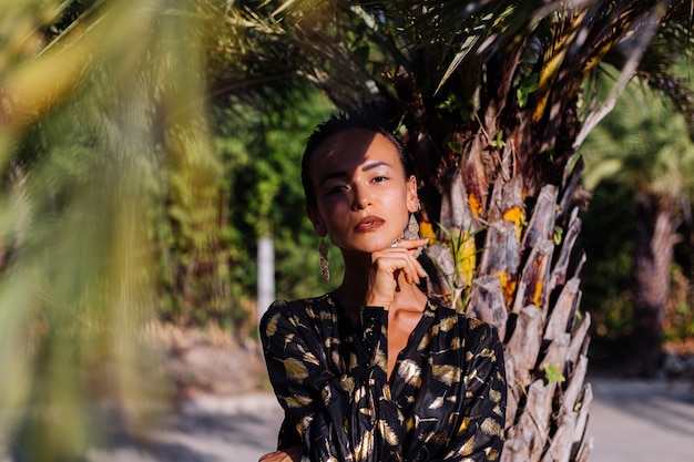 Mujer con maquillaje de bronce en vestido negro dorado cerca de una palmera