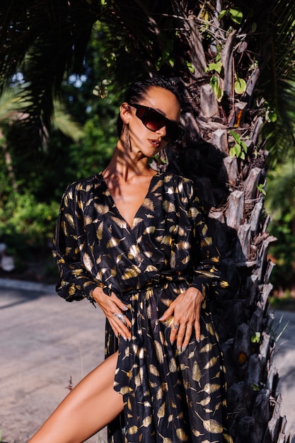 Mujer con maquillaje de bronce vistiendo un vestido negro dorado y gafas de sol en el paisaje tropical