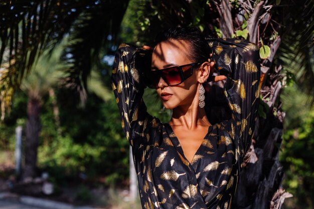 Mujer con maquillaje de bronce vistiendo un vestido negro dorado y gafas de sol en el paisaje tropical