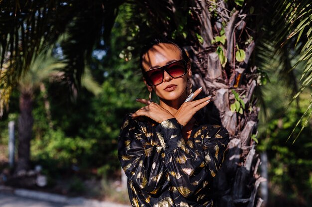 Mujer con maquillaje de bronce vistiendo un vestido negro dorado y gafas de sol en el paisaje tropical