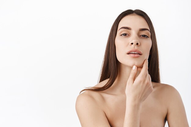 Mujer con maquillaje natural y retrato de piel sana. Modelo femenino hermoso que toca la piel facial hidratada que brilla intensamente fresca en el primer de la pared blanca. Concepto de cuidado de la piel