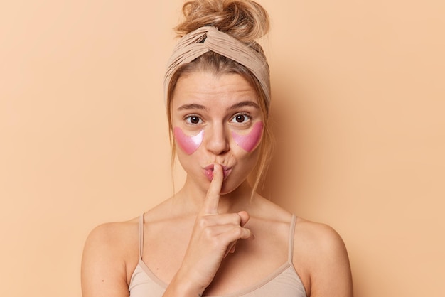 La mujer maravillada hace el signo de silencio, presiona el dedo índice sobre los labios, aplica parches de colágeno debajo de los ojos para reducir las arrugas o la hinchazón, usa una camiseta con banda para la cabeza aislada sobre un fondo beige. concepto de belleza