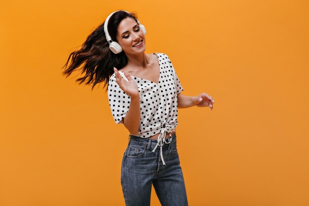 Mujer maravillosa baila y escucha música en auriculares blancos. Chica de pelo oscuro en blusa de lunares y jeans azul está sonriendo.