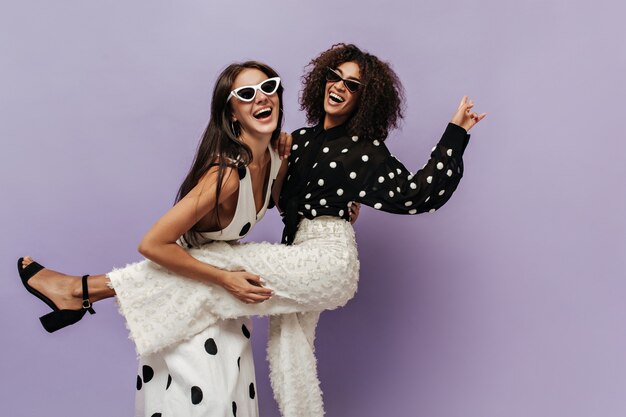 Mujer maravillosa en blusa negra y pantalones blancos de moda riendo y divirtiéndose con su amiga en gafas de sol en la pared aislada