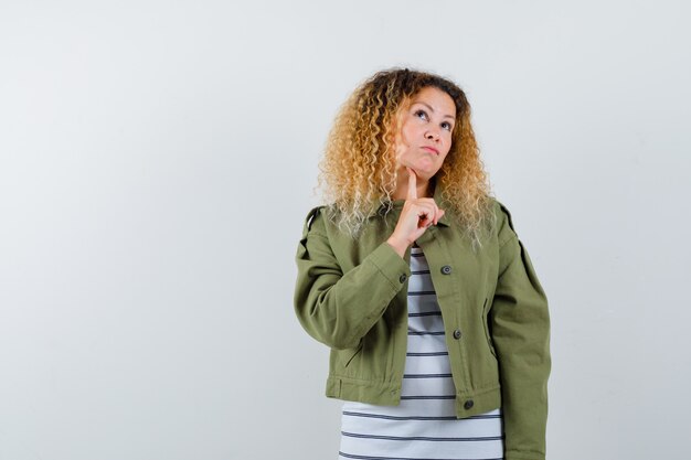 Mujer maravillosa en chaqueta verde, camisa manteniendo el dedo debajo de la barbilla, mirando hacia arriba y mirando pensativo, vista frontal.