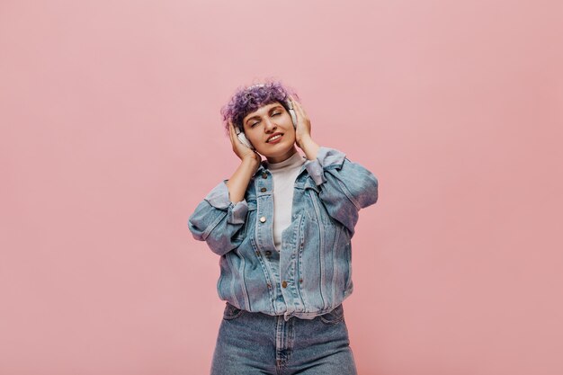 Mujer maravillosa en jeans frescos con cabello rizado lila disfruta de la música. Linda mujer en auriculares blancos posando.