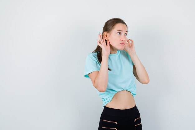 Mujer maravillosa manteniendo las manos detrás de las orejas en blusa, pantalones y mirando enfocado. vista frontal.