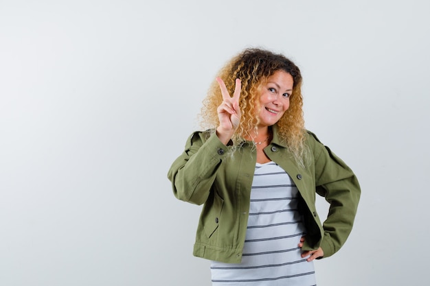 Mujer maravillosa que muestra el signo de V en chaqueta verde, camisa y mirando alegre. vista frontal.