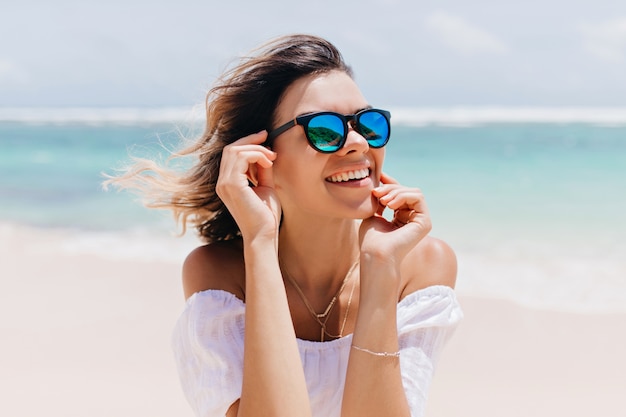 Mujer maravillosa en traje blanco y gafas brillantes posando con expresión de cara feliz en un caluroso día de verano. agradable mujer caucásica de pie cerca del océano en el cielo