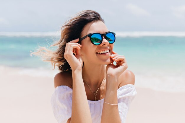 Mujer maravillosa en traje blanco y gafas brillantes posando con expresión de cara feliz en un caluroso día de verano. Agradable mujer caucásica de pie cerca del océano en el cielo