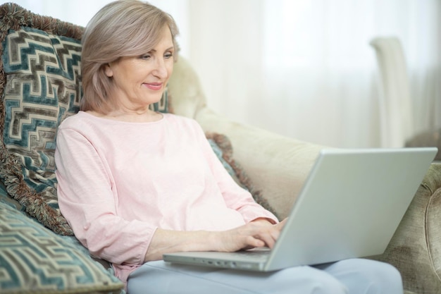 Mujer de más de 50 años se sienta frente a una computadora portátil en casa. Escribe una carta a su hija. La mujer tiene una sonrisa tierna en el rostro.
