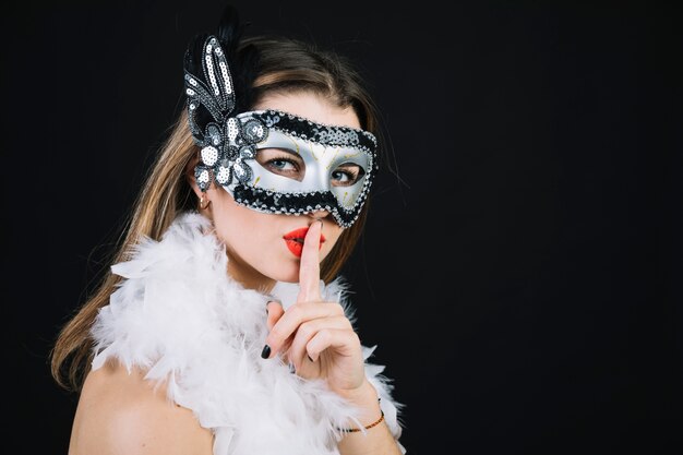 Mujer con una máscara de carnaval haciendo gesto de silencio sobre fondo negro