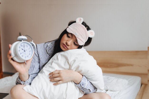 Mujer en máscara de dormir rosa está abrazando la almohada y mirando el despertador con tristeza