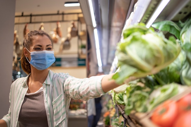 Mujer con máscara higiénica y guantes de goma y carrito de compras en el supermercado comprando verduras durante el virus corona y preparándose para una cuarentena pandémica