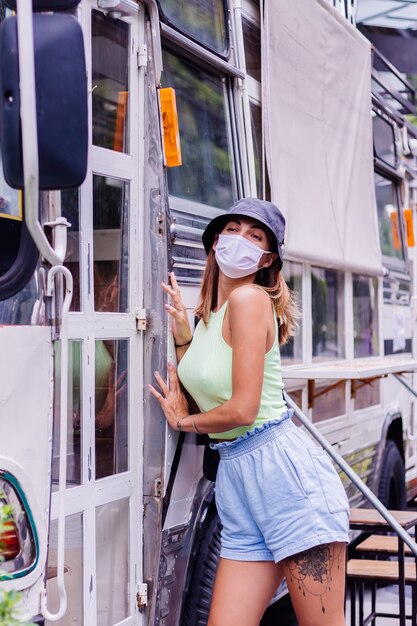 Mujer en máscara médica blanca caminar por la ciudad se encuentra en la cafetería del autobús en la plaza de la ciudad
