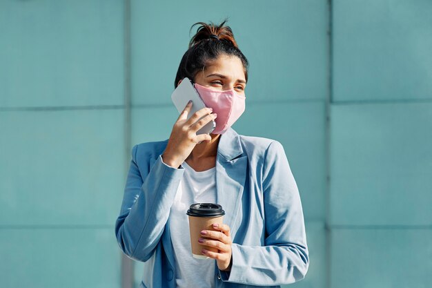 Mujer con máscara médica y café hablando por teléfono inteligente en el aeropuerto durante la pandemia