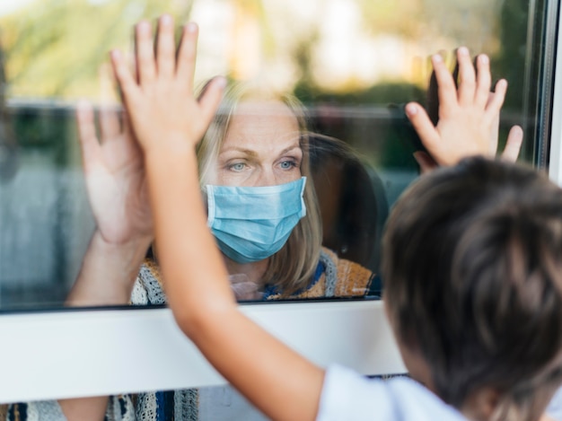 Mujer con máscara médica en casa saludando a su sobrino a través de la ventana