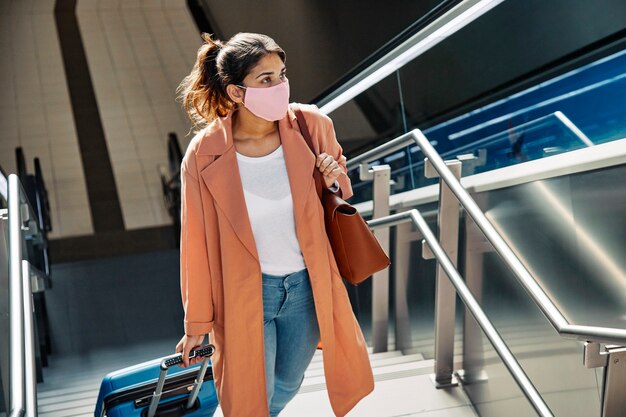 Mujer con máscara médica y equipaje subiendo escaleras en el aeropuerto durante la pandemia