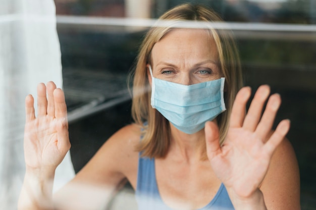 Mujer con máscara médica mirando por la ventana durante la cuarentena