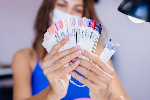 Mujer con máscara médica protectora en el salón de belleza, sostenga la paleta y seleccione un color Procedimiento de manicura Cuidado de las uñas Gran inauguración La cuarentena ha terminado Las pequeñas empresas están abiertas nuevamente