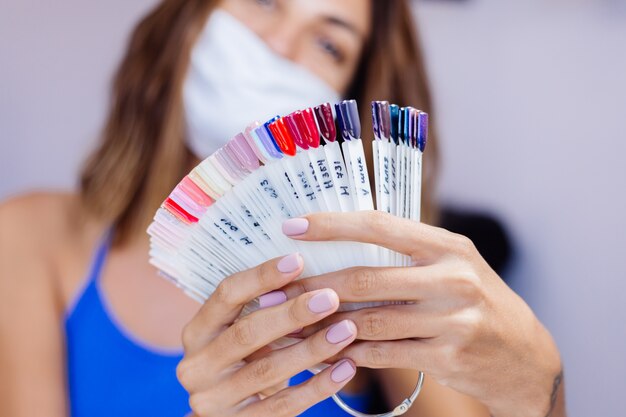 Mujer con máscara médica protectora en el salón de belleza, sostenga la paleta y seleccione un color Procedimiento de manicura Cuidado de las uñas Gran inauguración La cuarentena ha terminado Las pequeñas empresas están abiertas nuevamente