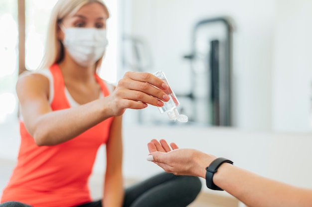 Mujer con máscara médica que ofrece desinfectante de manos a la persona en el gimnasio