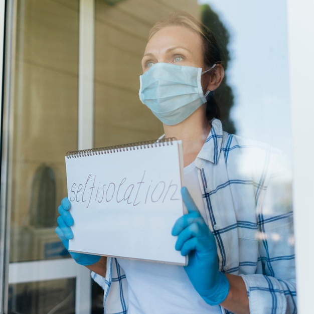 Foto gratuita mujer con máscara médica sosteniendo el cuaderno en casa en la ventana