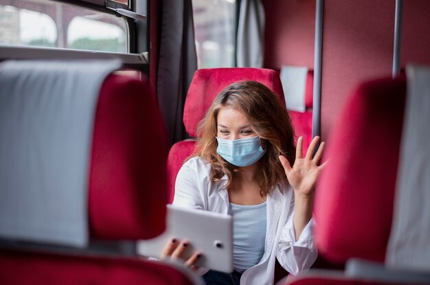 Mujer con máscara médica con tableta para videollamadas mientras viaja en tren público