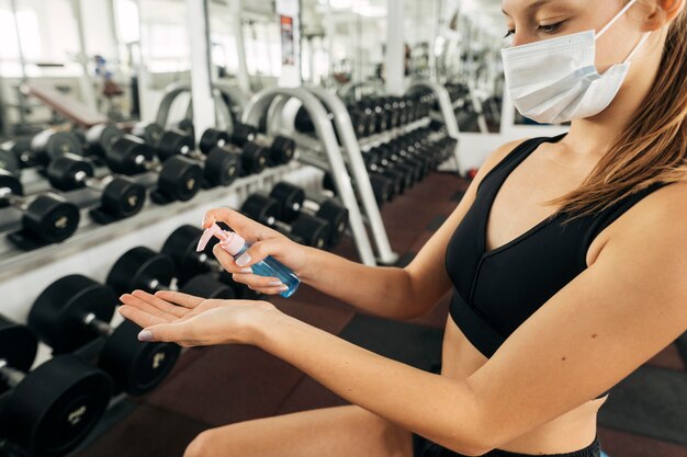 Mujer con máscara médica trabajando en el gimnasio y usando desinfectante para manos