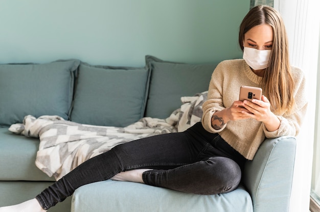 Mujer con máscara médica usando su teléfono inteligente en casa durante la pandemia