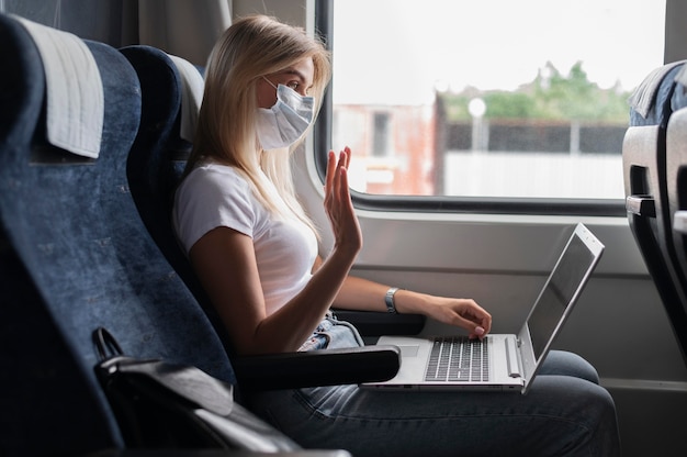 Mujer con máscara médica viajando en tren público y teniendo una videollamada en la computadora portátil