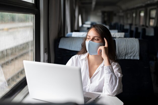 Mujer con máscara médica viajando en tren público y usando laptop