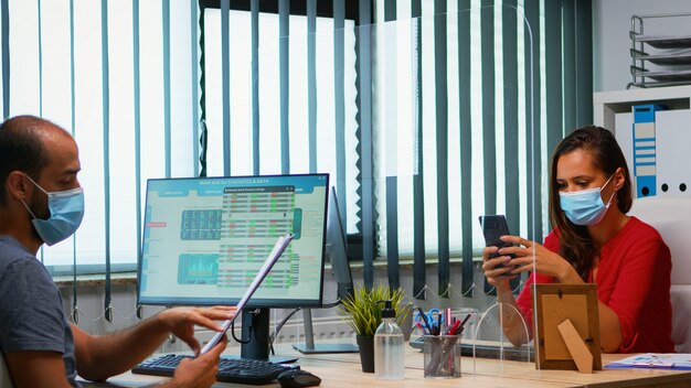 Mujer con máscara protectora escribiendo en el teléfono mientras su colega trabaja con portapapeles respetando el distanciamiento social. Freelancer en la nueva oficina normal charlando escribiendo en el móvil con tecnología de internet