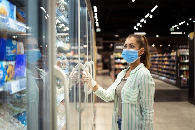 Mujer con máscara protectora y guantes abriendo congelador en supermercado durante la pandemia COVID-19 o el virus corona